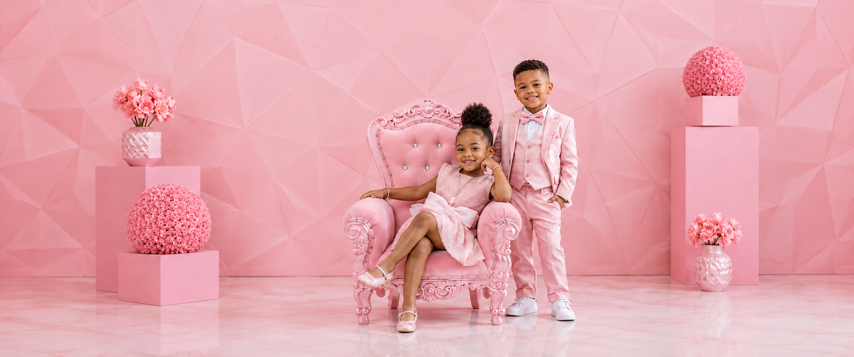 Two children dressed in pink pose with a pink royal mini throne chair in an elegant pink floral studio setting.