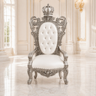 A royal-style throne chair with an ornate silver carved frame, a tufted white backrest, and a white cushioned seat, set in a bright classical interior with large windows and marble floors.