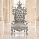 A royal-style throne chair with ornate silver carved woodwork, a tufted backrest, and a crown on top, set in an elegant cream-colored palace interior with marble floors and chandeliers.