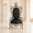 A luxurious throne-style armchair with an ornate silver carved frame, a black tufted leather backrest, and a crown topper, set in a grand, elegant room with cream walls and marble floors.