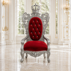 An ornate royal-style throne chair with a silver carved frame and a deep red velvet seat and backrest, set in a bright classical hall with large windows and polished marble floors.