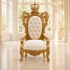 An ornate royal-style armchair with a carved gold frame and a tufted cream upholstered backrest, topped with a crown motif, set in a bright, classical interior with large windows and polished floors.