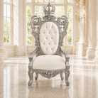 An ornate royal-style throne chair with a silver carved frame, tufted white backrest, and white seat cushion, placed in a bright classical interior with large windows and columns.
