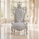 An ornate silver throne chair with a tufted backrest and crown detail, set in a bright, classical room with marble floors and large windows.