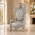A highly ornate silver baroque-style throne chair with a tufted backrest and a crown on the backrest, set in a luxurious cream-colored room with marble floors.