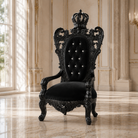 An ornate black throne chair with a high carved back, crown topper, tufted button detailing, and carved arms, set in a luxurious beige room with marble floors and classical columns.