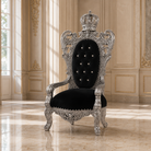 An ornate royal throne chair with a silver carved frame, crown on the backrest, and black tufted upholstery, set in a luxurious cream-colored room with gold paneling and marble floors.