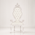 A white baroque-style high-back vanity chair with tufted upholstery, ornate carved details, and silver nailhead accents, set against a white paneled wall background.